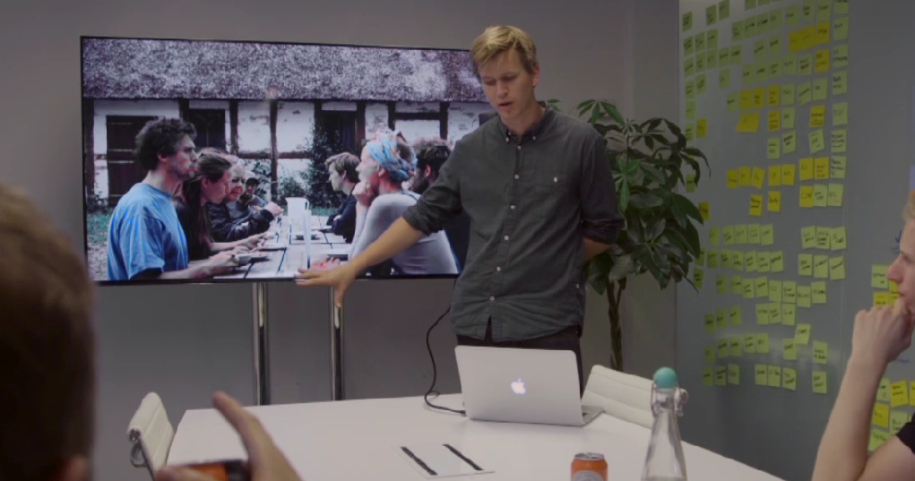 Skjermdump fra NRK skole serien. Mann som står foran en tv og folk rundt et bord.